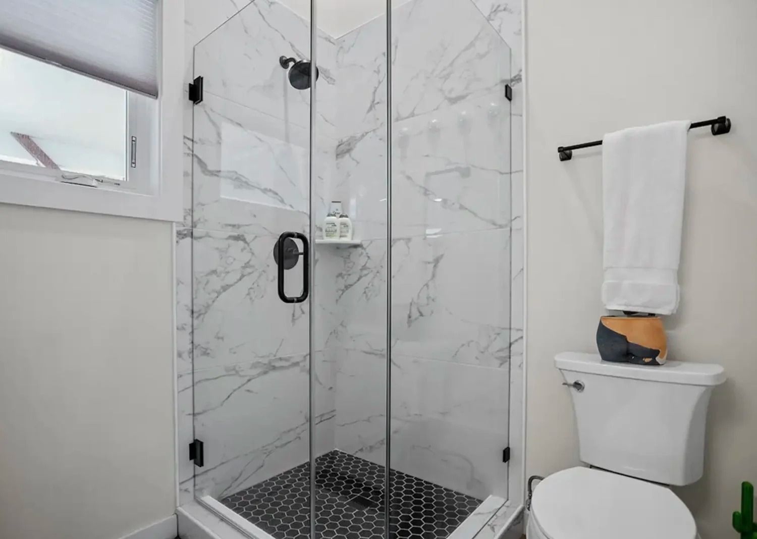 Bright bathroom with glass shower, black fixtures, white marble walls, and a toilet with a towel on top