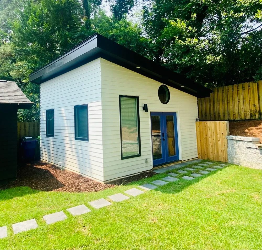 Small white garden shed with black roof, blue door, and stepping-stone path in a sunny backyard