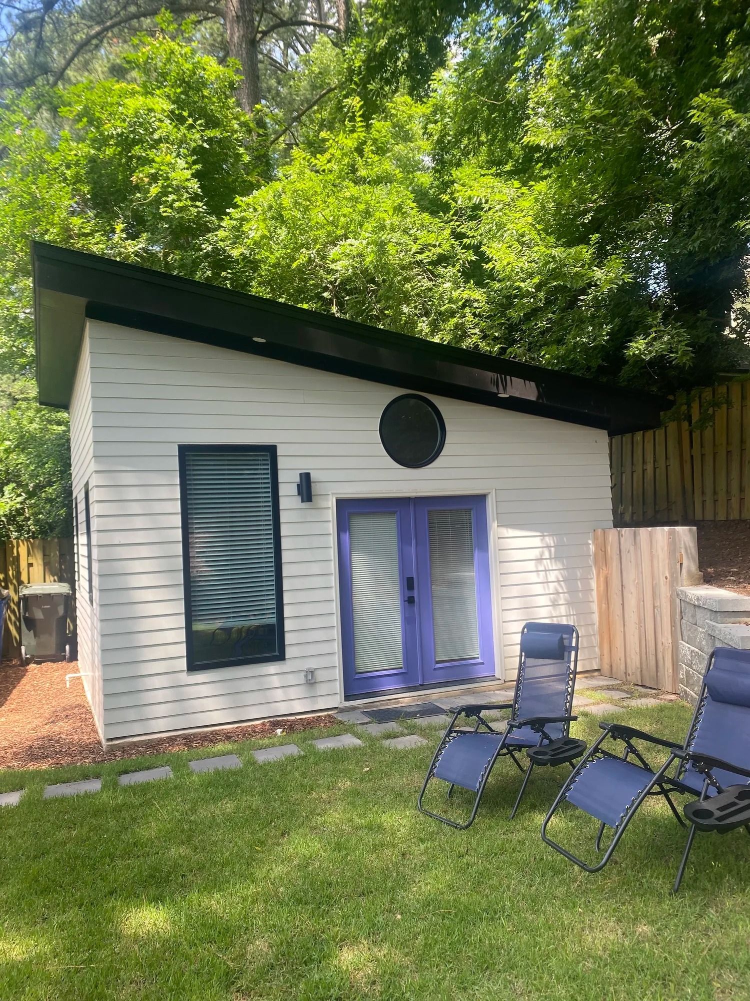 Small white cottage with black roof and purple doors, set in a grassy yard with folding chairs and trees.