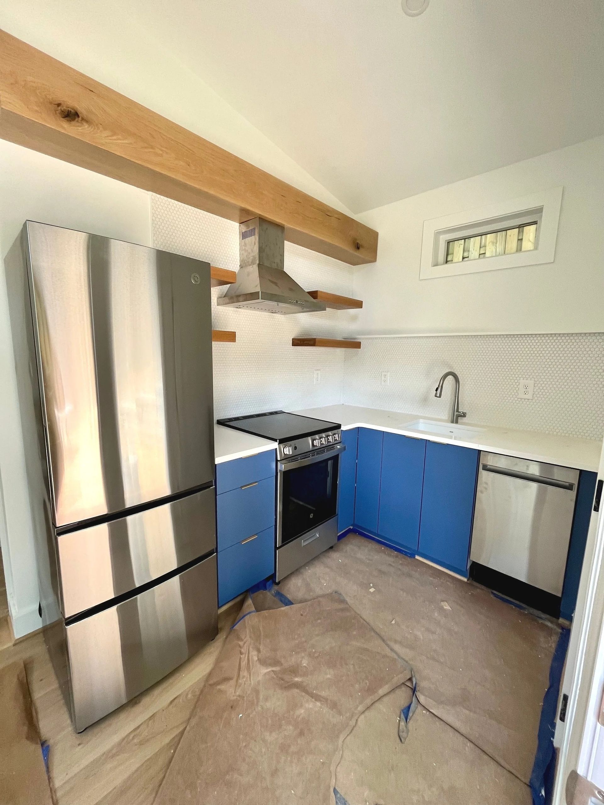 Small kitchen with stainless fridge, blue cabinets, stove, sink, and exposed wooden beam ceiling.
