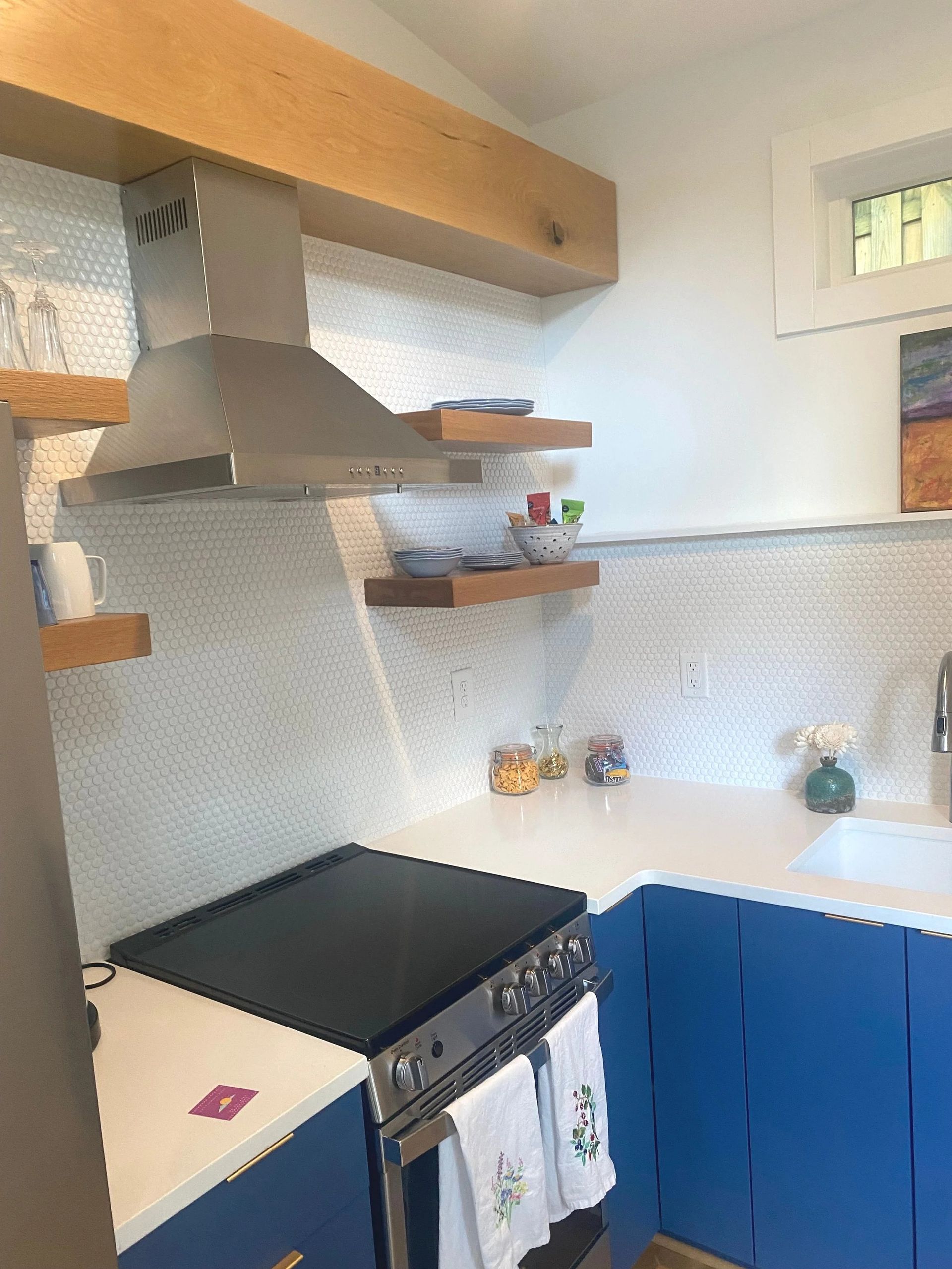 Small kitchen with blue cabinets, stove, wooden shelves, and a window above a sink area.