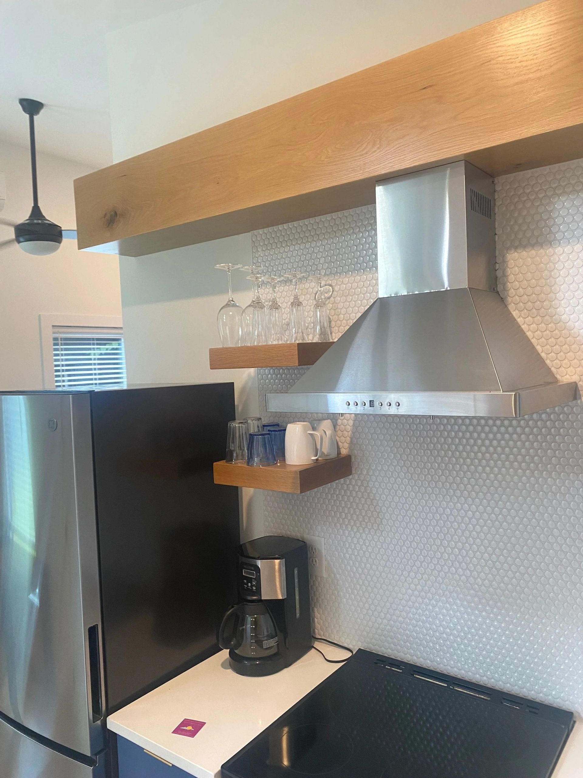 Modern kitchen corner with stainless steel range hood, black fridge, coffee maker, and hanging glasses