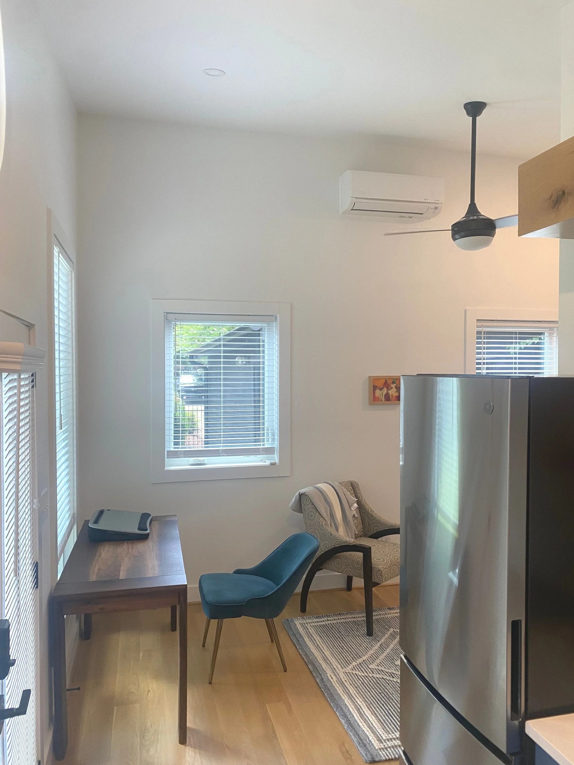 Small living space with a sofa, desk, chair, rug, ceiling fan, and refrigerator by a window.