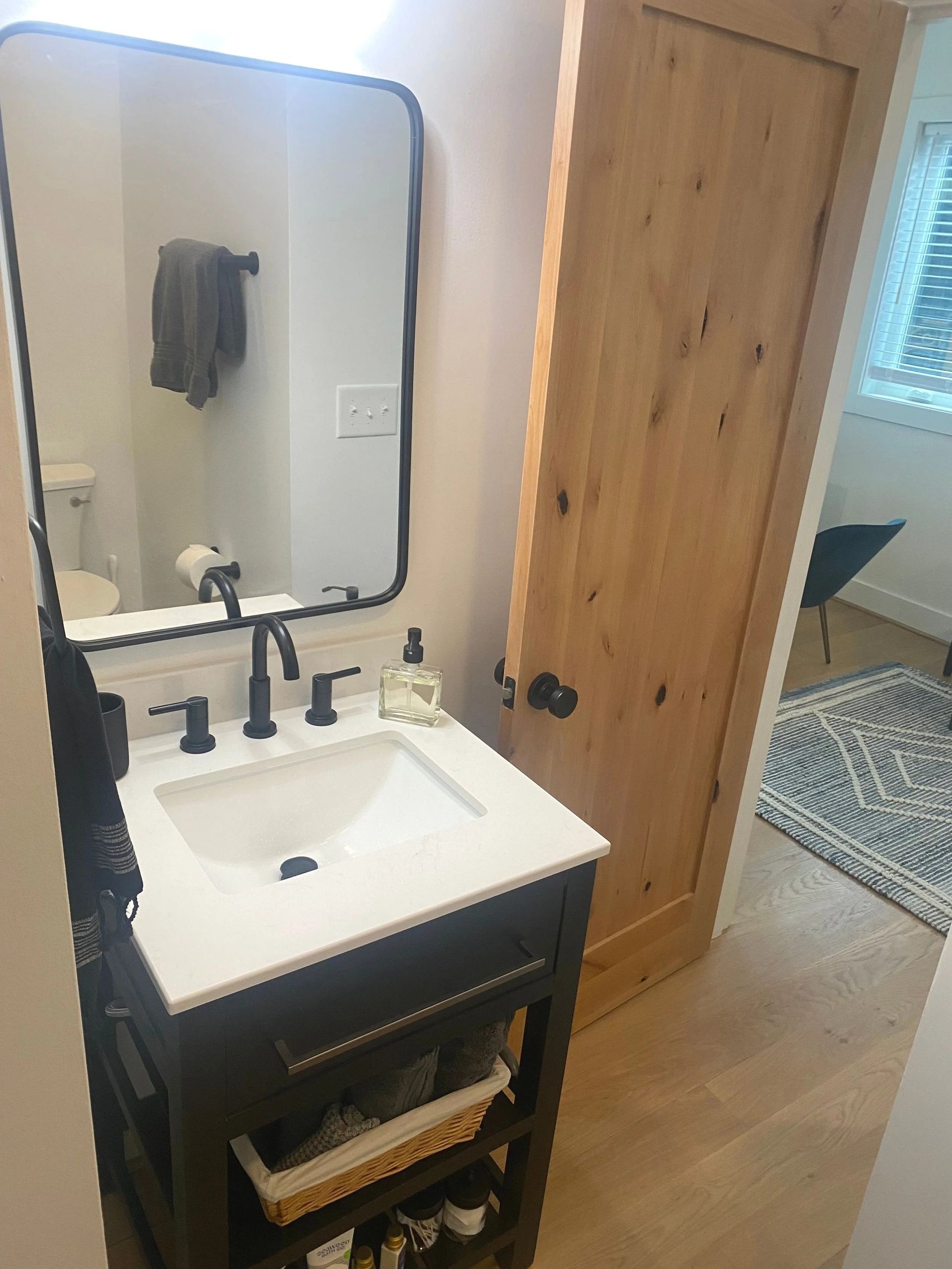 Small bathroom vanity with mirror, black faucet, soap dispenser, and wood door beside a bedroom.