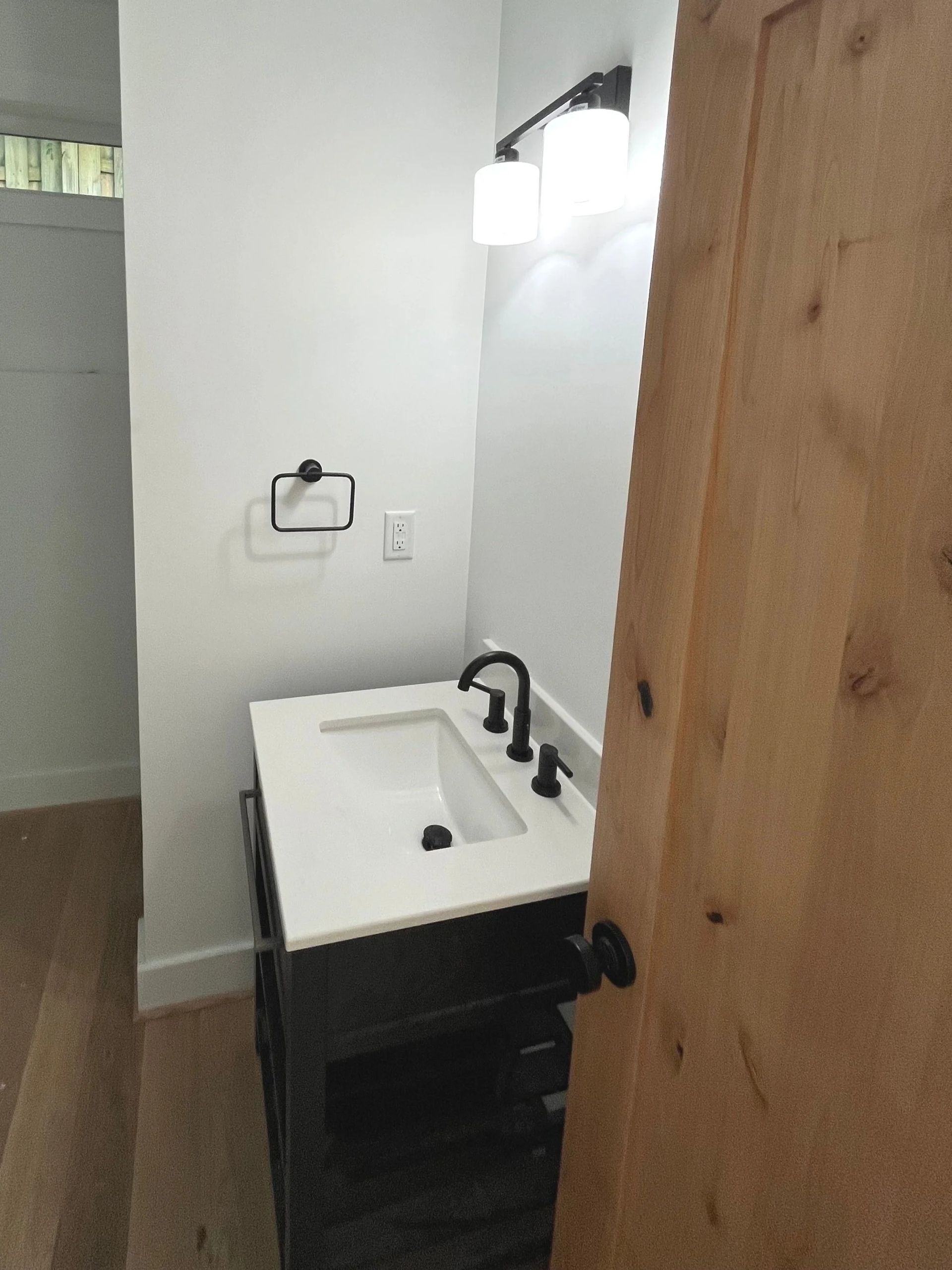 Small bathroom vanity with white sink, black faucet, wall light, and wood door.