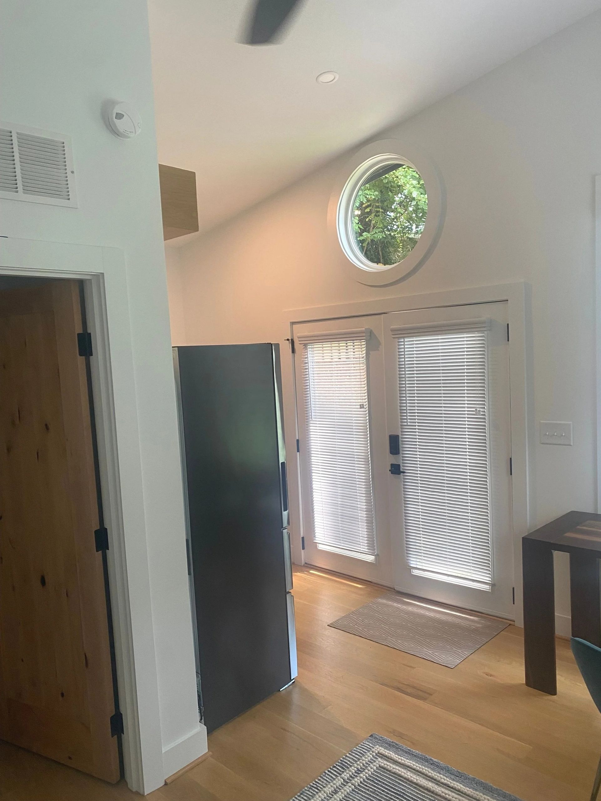 Bright entryway with white double doors, round window, wood floor, and tall black cabinet beside a wooden front door