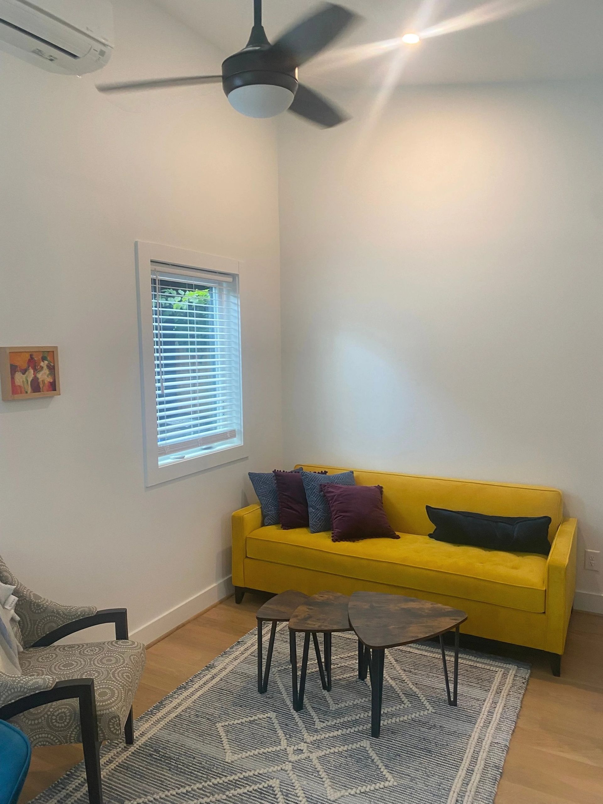 Bright living room with yellow sofa, patterned rug, small tables, ceiling fan, and window blinds