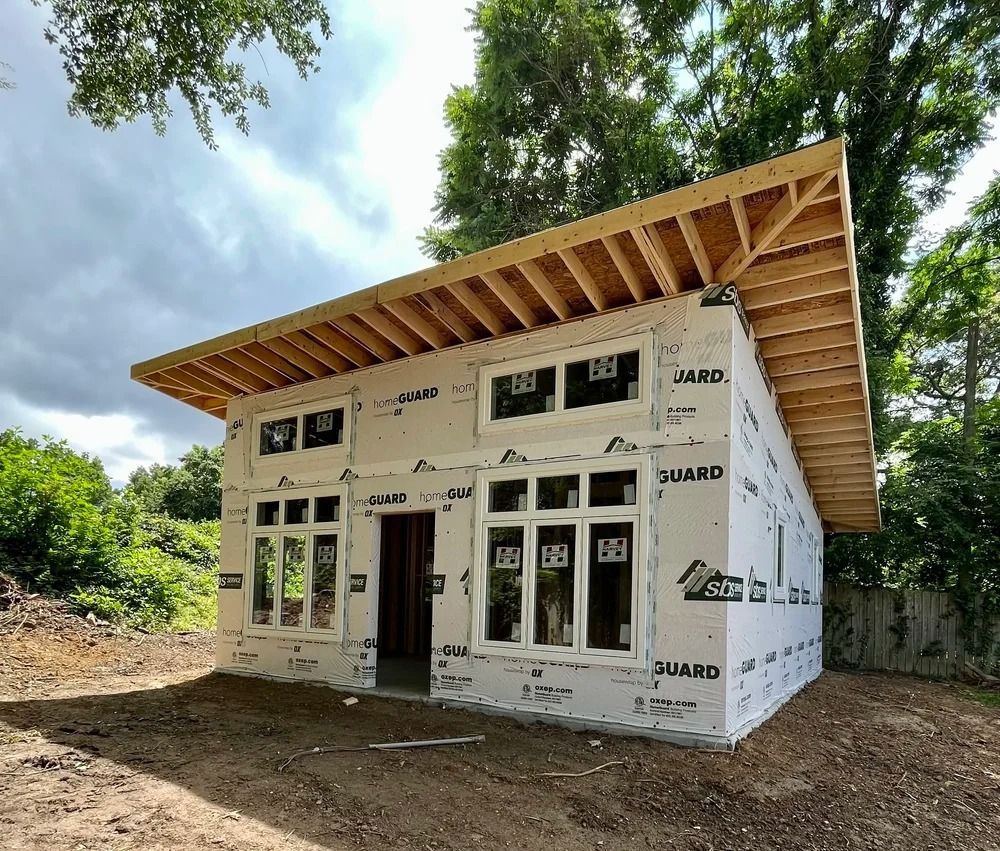 Small unfinished house under construction with white exterior wrap and exposed wooden roof framing in a wooded lot