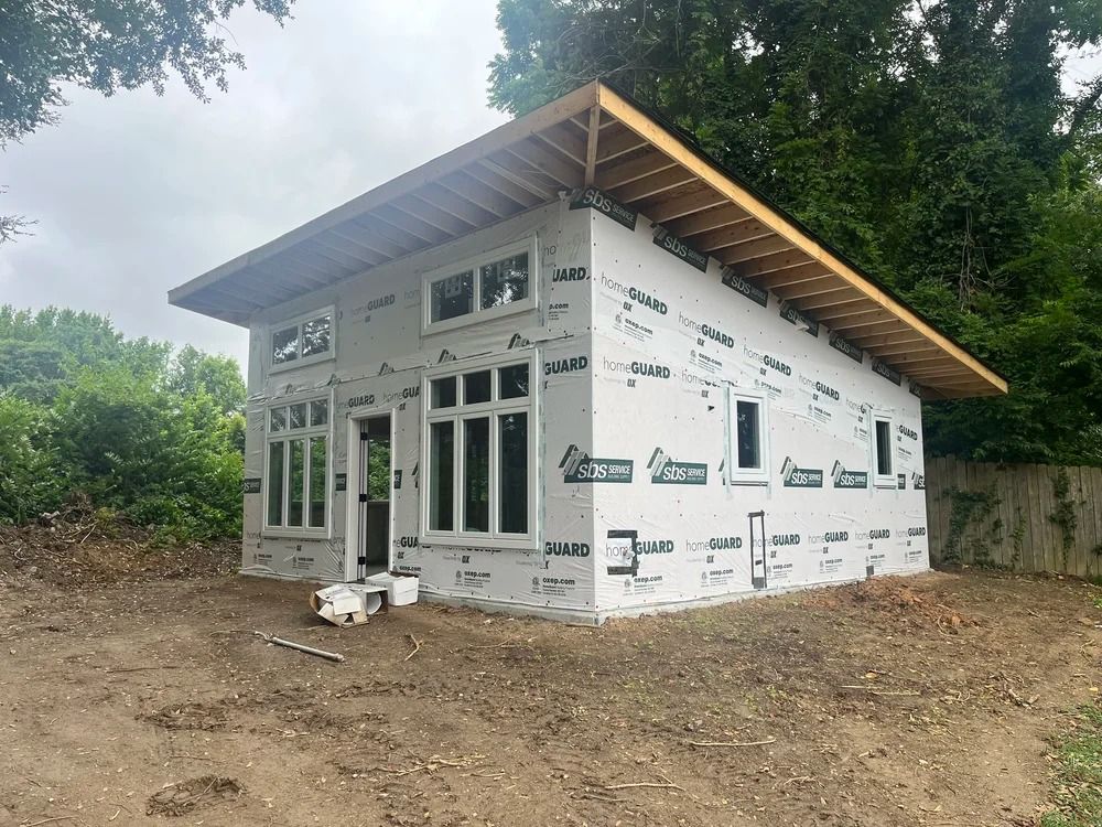 Small unfinished white house under construction on a dirt lot, surrounded by trees and a cloudy sky.
