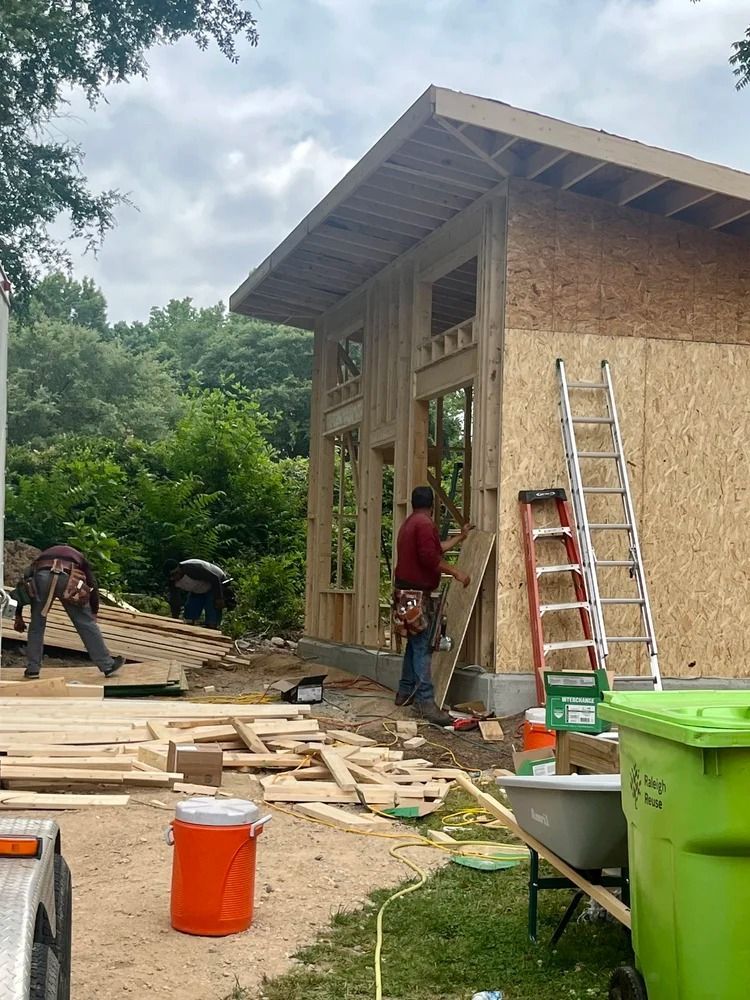 Workers building an unfinished house exterior with ladders, wood planks, and a green bin