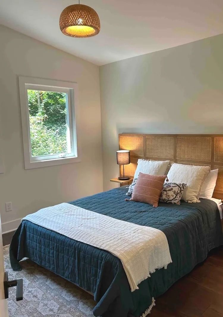 Small bedroom with a bed, teal quilt, woven headboard, lamp, and window with trees outside