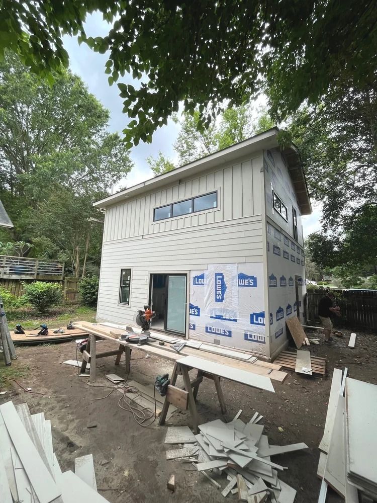 Small white house under construction with workers, scaffolding, and building materials in a wooded yard