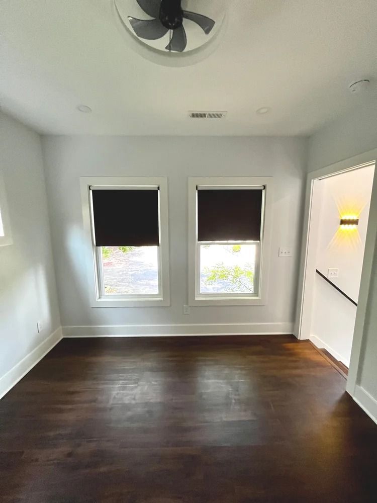 Empty white room with dark wood floors, two windows with black blinds, and a ceiling fan