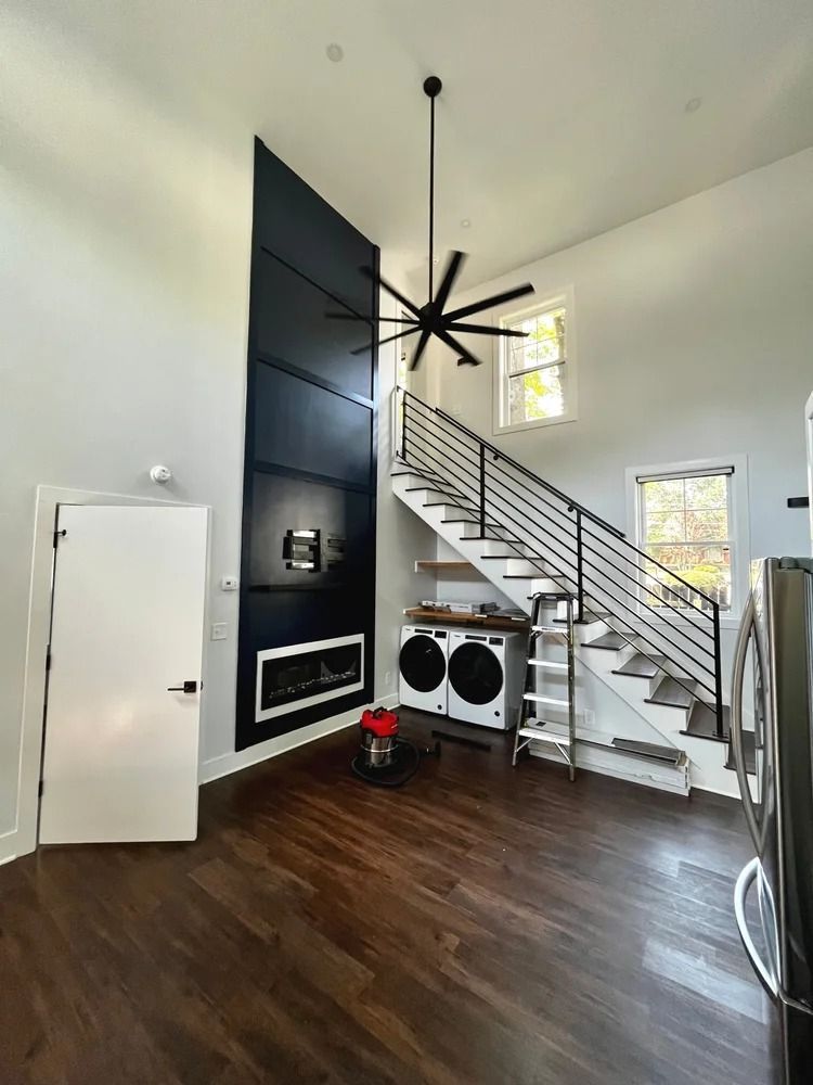 Modern living room with dark wood floors, a black accent wall, staircase, and large ceiling fan
