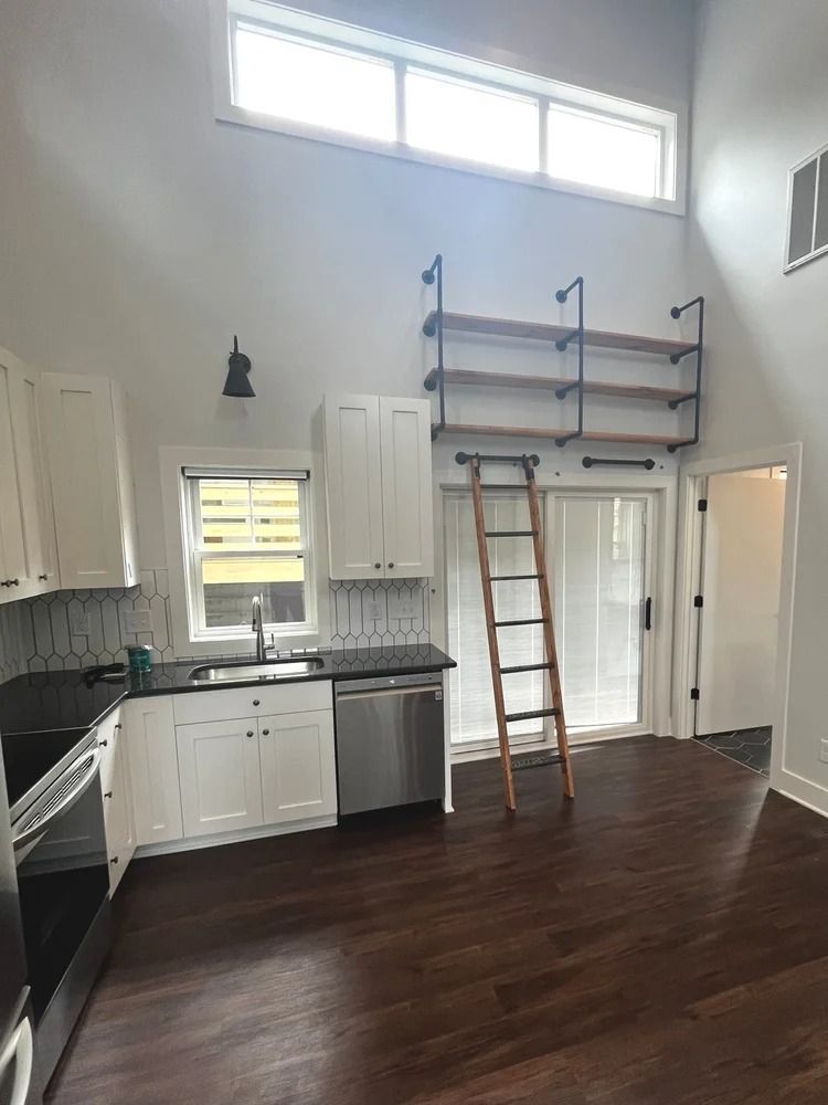 Empty kitchen with dark wood floors, white cabinets, high windows, and a ladder leaning against built-in shelving.