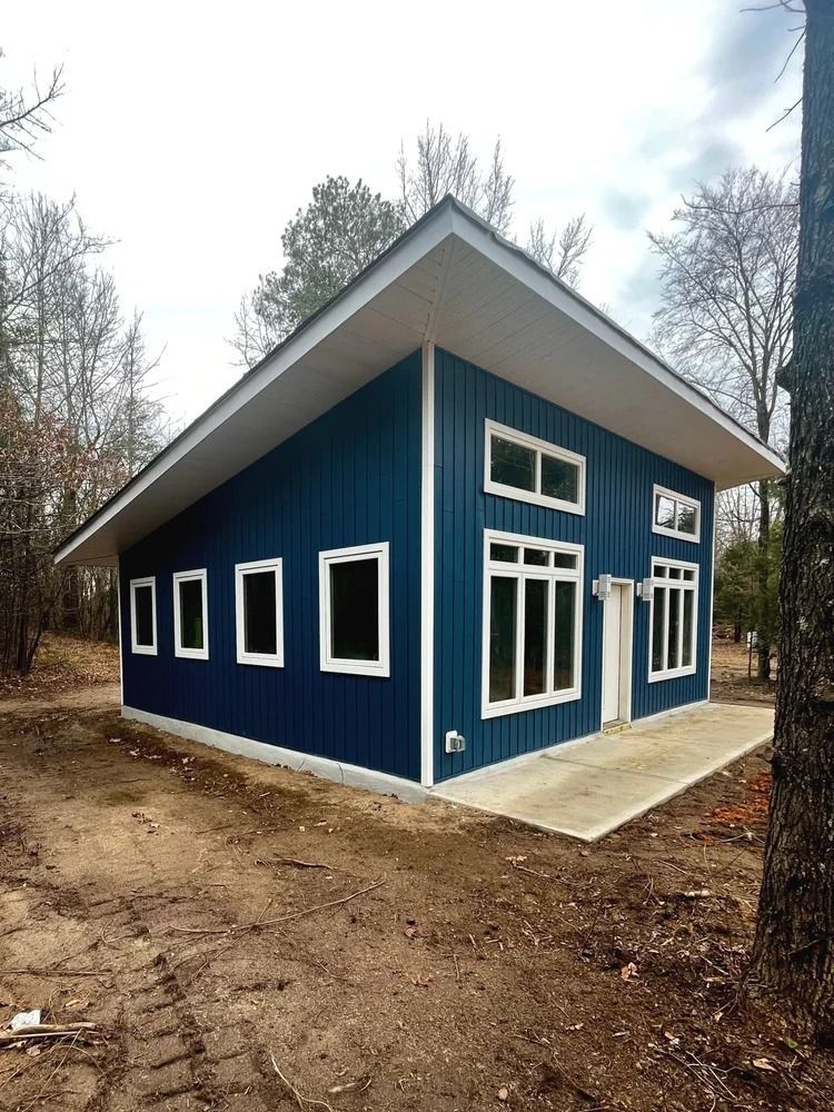 Blue modern cabin with white trim and sloped roof on a concrete pad in a wooded area