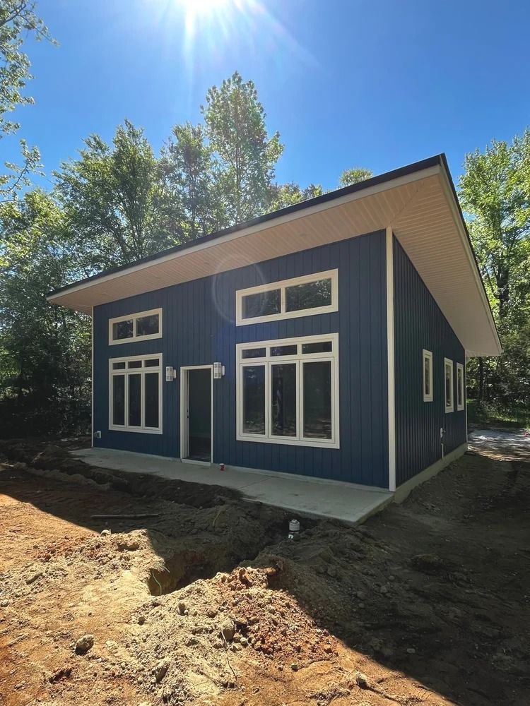 Blue house with white trim under construction on a sunny lot, surrounded by dirt and trees