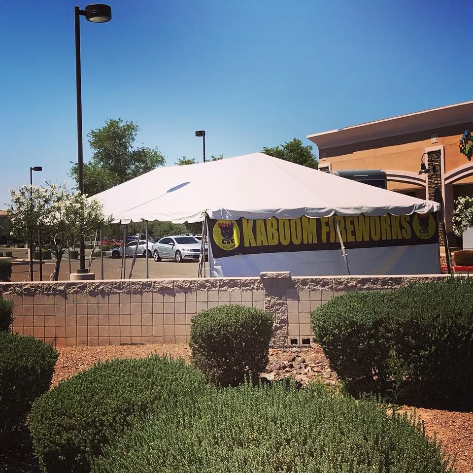 White event tent with a black-and-gold banner in a sunny parking lot beside a brick building.
