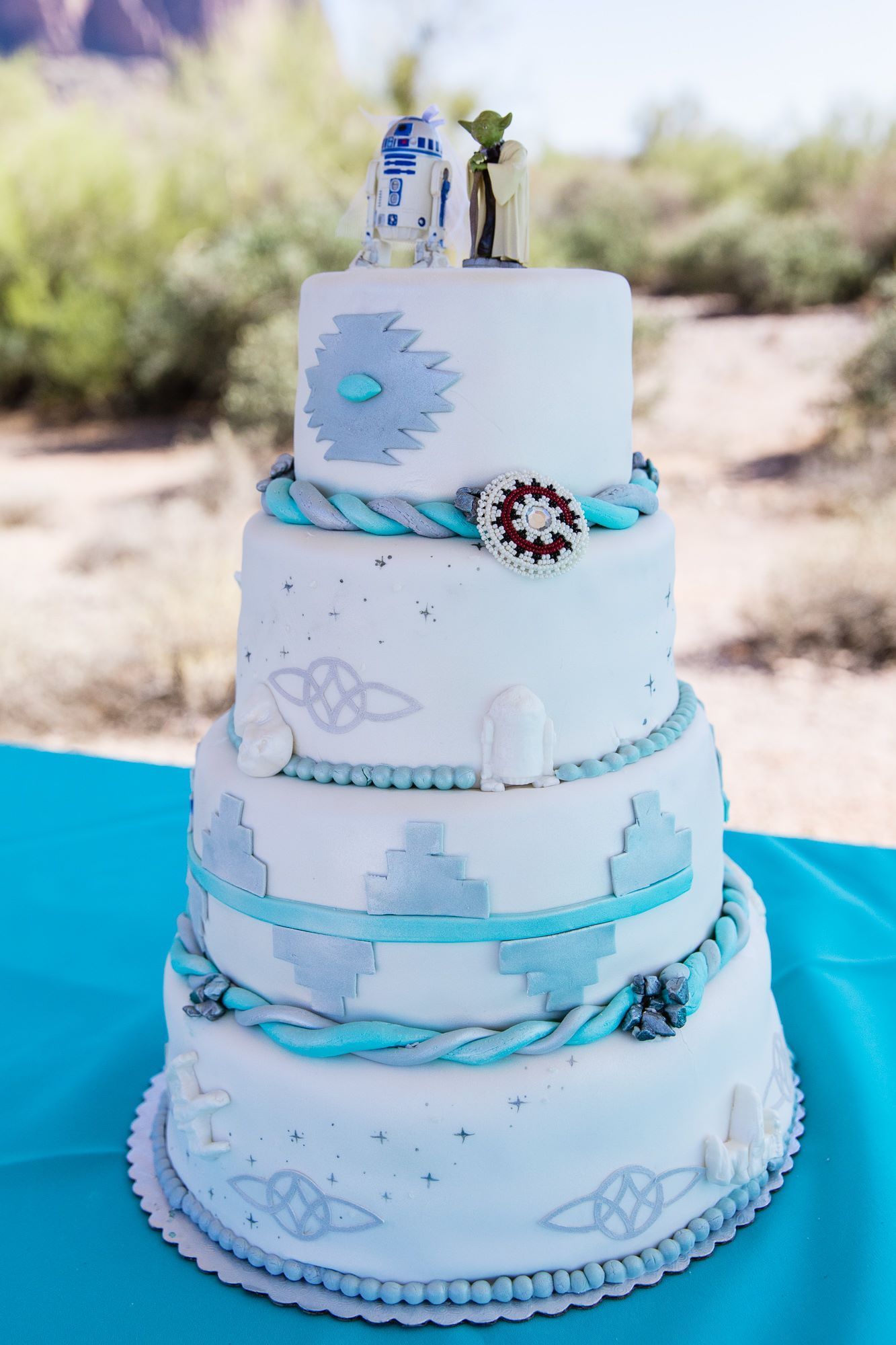 Four-tier white cake with blue ribbon accents and seashell decorations on a turquoise table outdoors