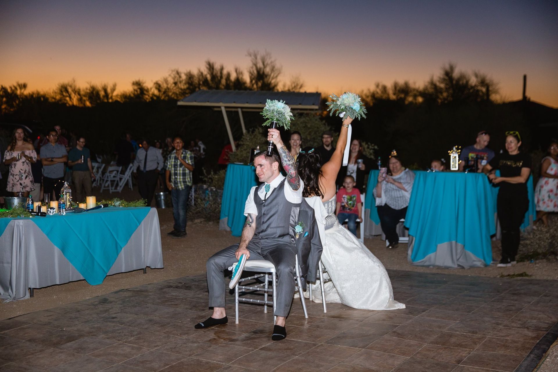 Bride and groom in a wheelchair and on a white chair, raising pom-poms at an outdoor sunset celebration