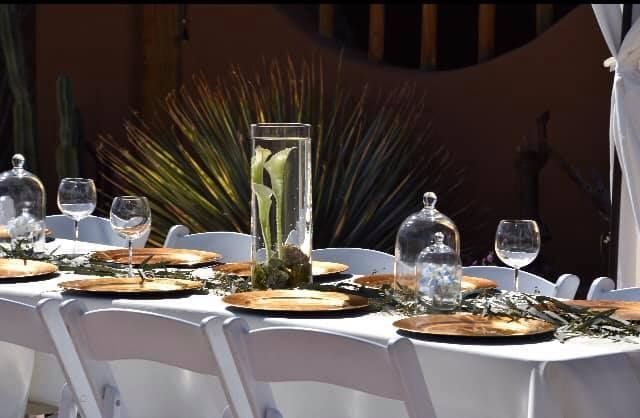 Elegant outdoor dinner table with white chairs, gold plates, glassware, and a tall floral centerpiece
