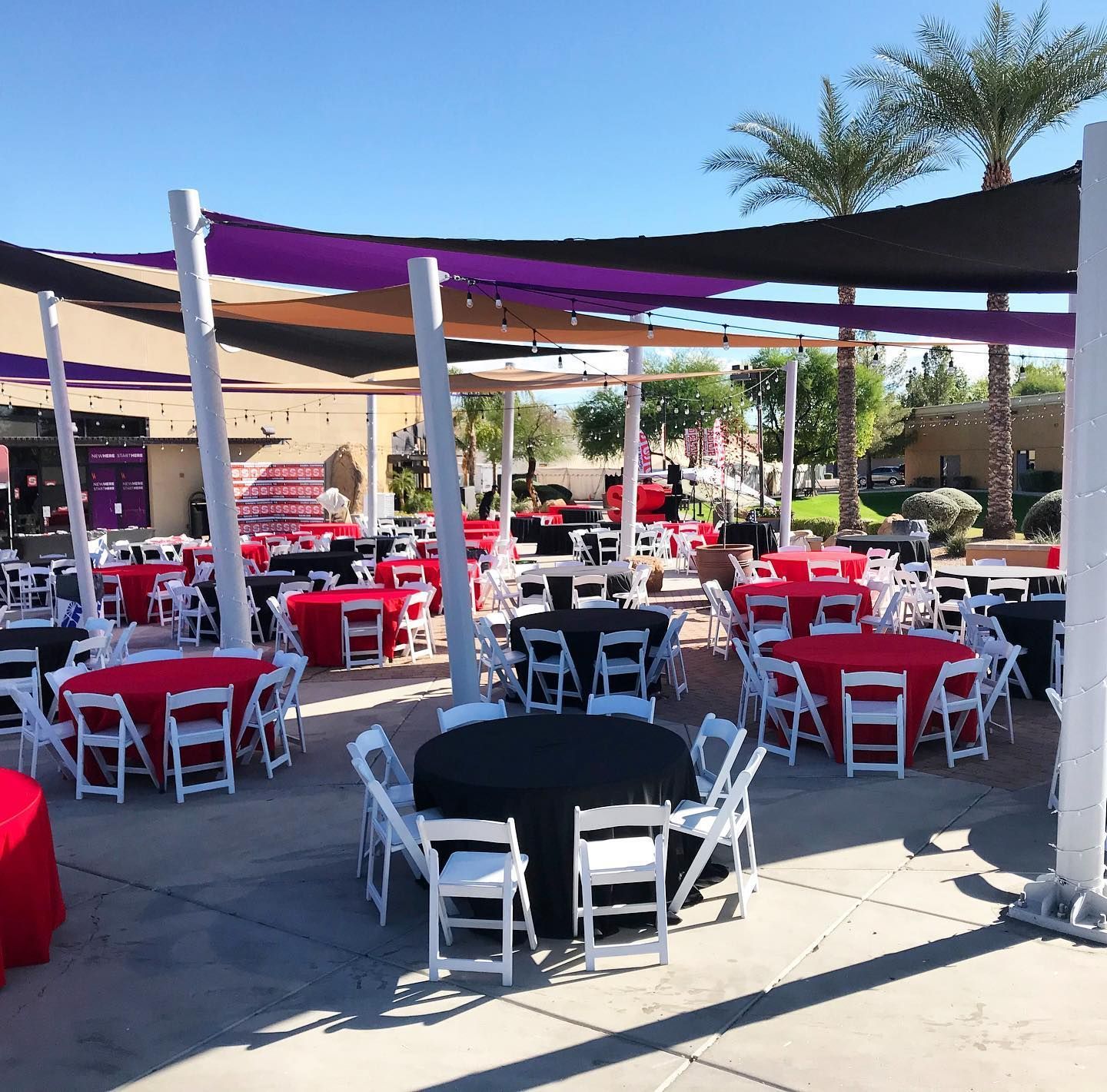 Outdoor patio dining area with red and white tables under purple shade sails on a sunny day