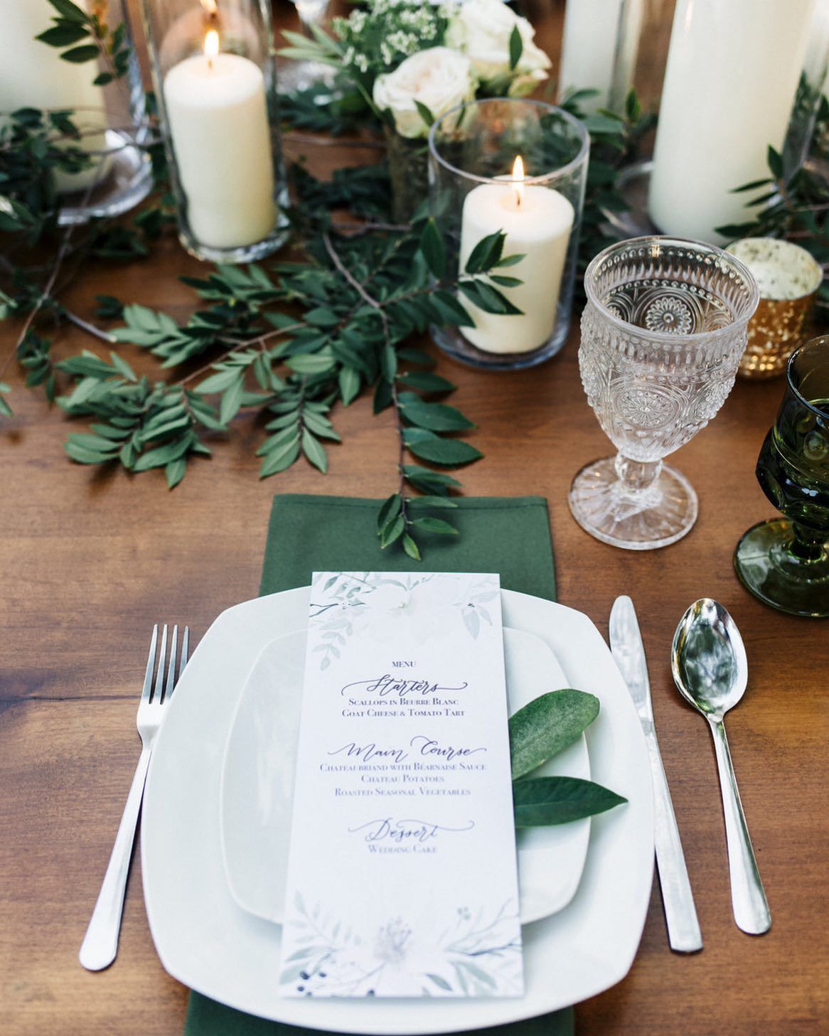 Elegant table setting with white candles, greenery, and a personalized place card on a white plate