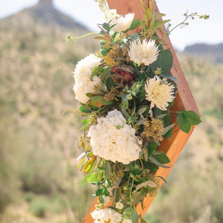White floral arrangement with greenery on a wooden frame outdoors, with blurred trees in the background