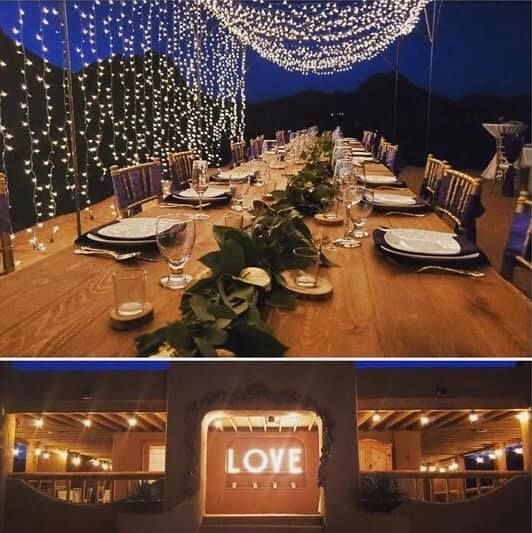 Long wooden dining table set under string lights, with a glowing “LOVE” sign in a rustic venue