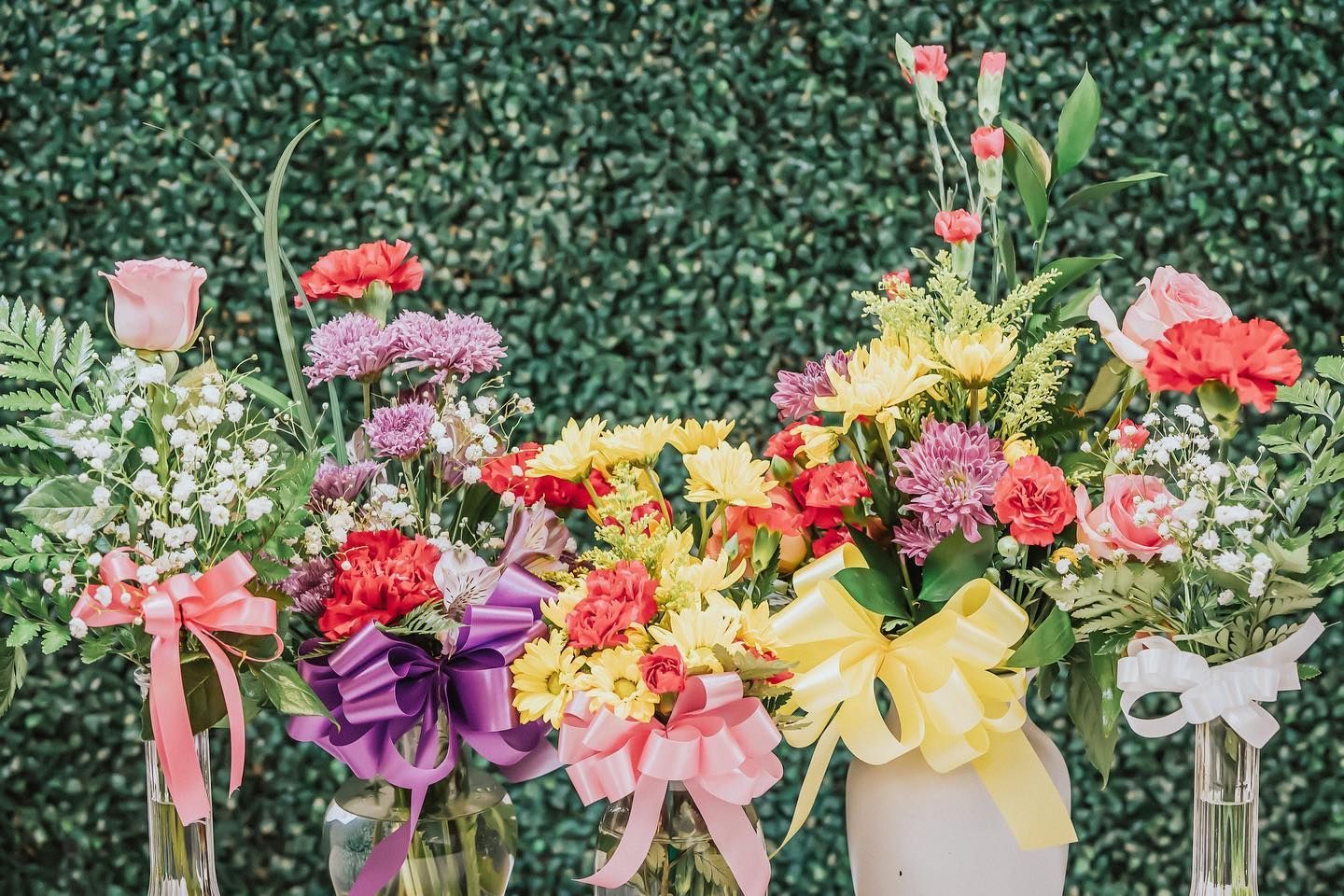 Colorful flower bouquets in vases with pink, purple, yellow, and red blooms against a green hedge backdrop
