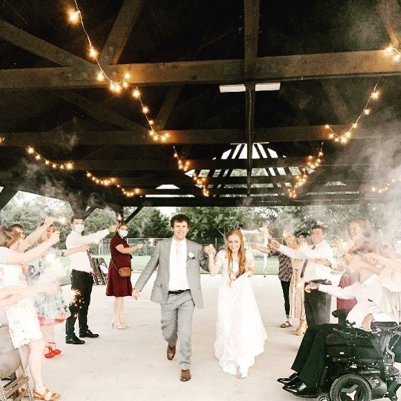 Newlyweds walking under a wooden pavilion with string lights and smoke, guests cheering around them