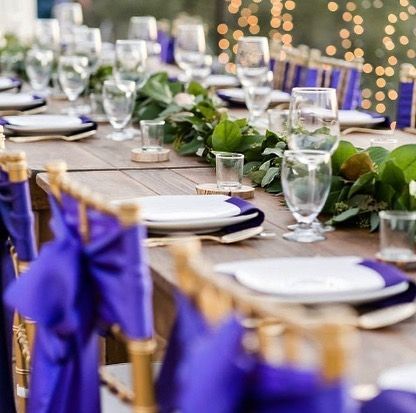 Elegant outdoor dining table with purple chair sashes, white plates, glassware, and greenery centerpiece