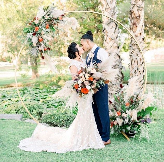 Wedding couple kissing under floral arch in a bright garden, bride in white gown and groom in navy suit