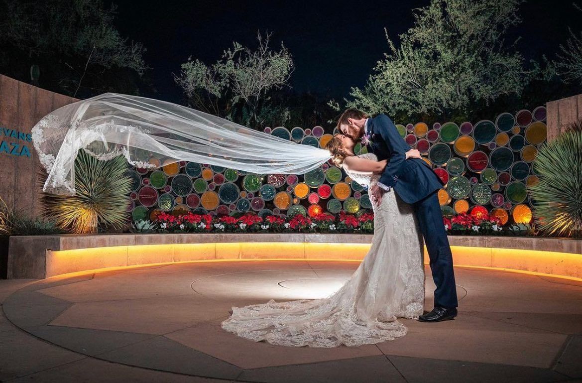 Bride and groom kissing as her veil billows in the wind on a lit outdoor terrace at night