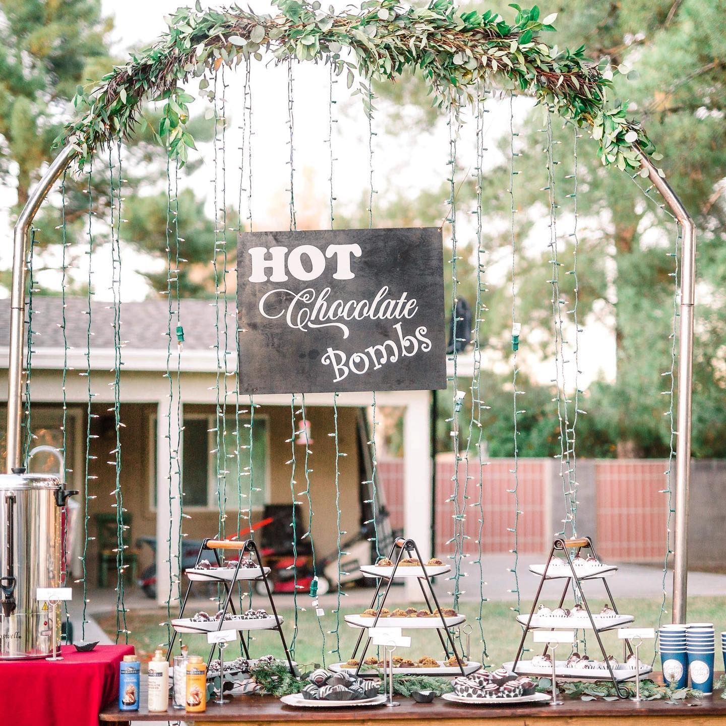 Outdoor hot chocolate bar under a floral arch with hanging lights and a “HOT Chocolate Bombs” sign
