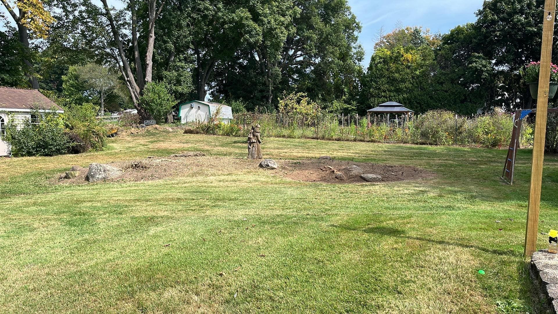 A grassy yard with a small garden area in the middle, trees and small buildings in the background.