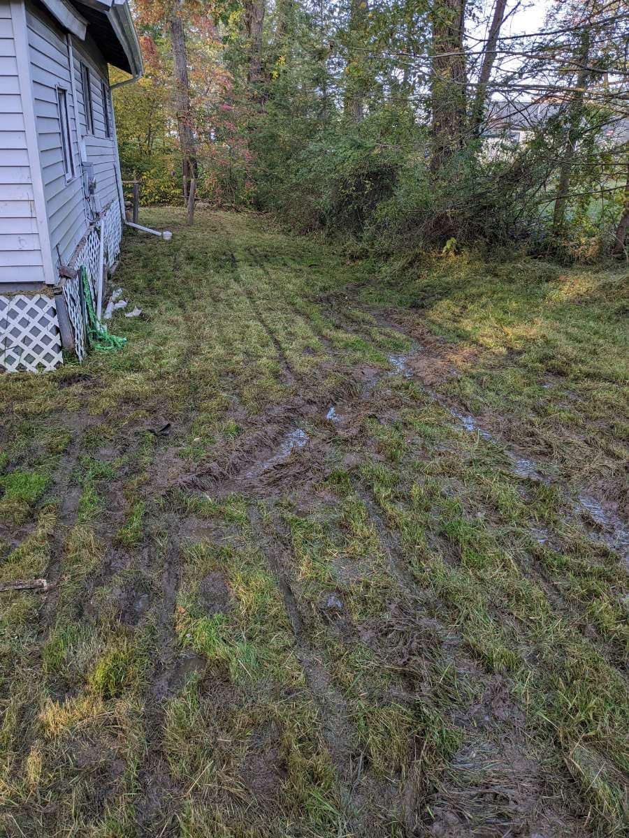 Muddy yard with tire tracks next to a light-colored house. Green grass and trees are in the background.