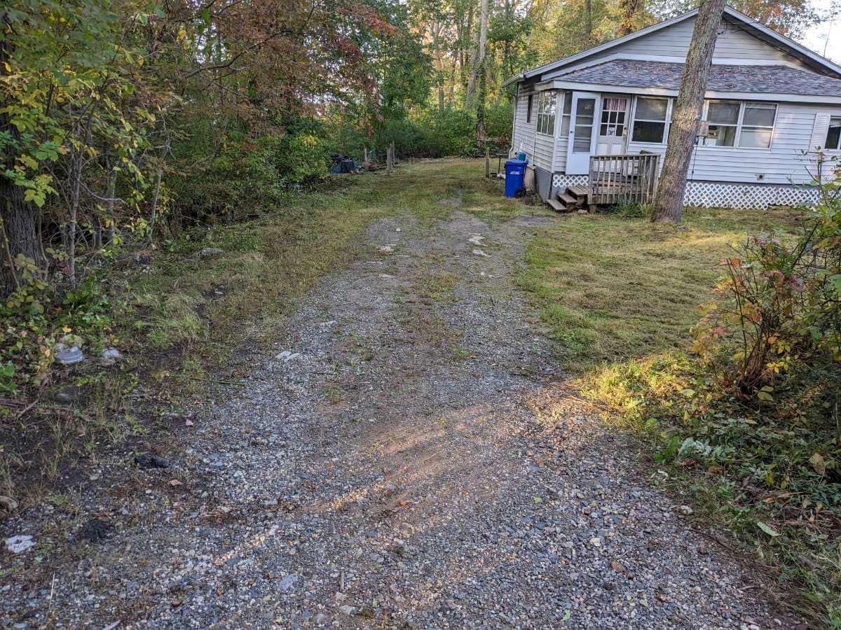 Gravel driveway leads to a weathered, white house. Vegetation borders the path.