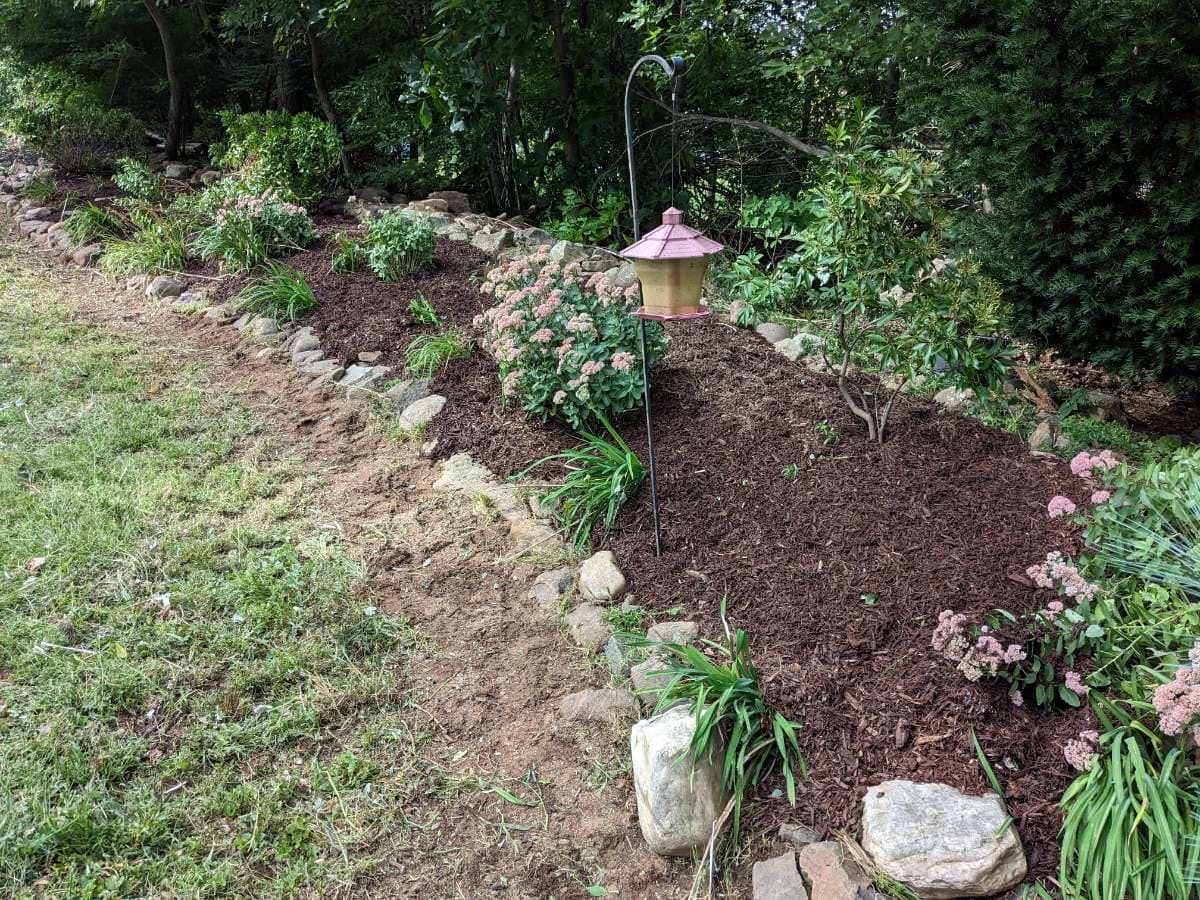 A landscaped garden bed with mulch, plants, and stone border; a decorative bird feeder is in the center.