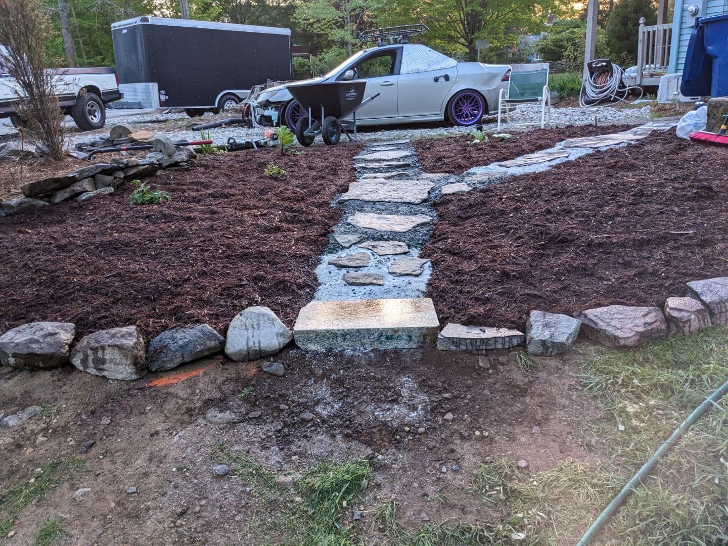 Stone path through a mulched garden bed, bordered by rocks, with a car and trailer in the background.