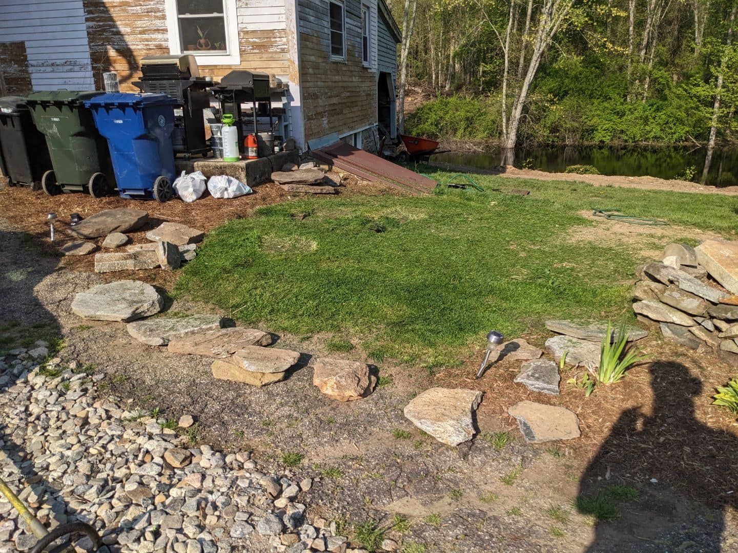 Yard with grass, rocks, and house. Green and blue trash cans are visible.