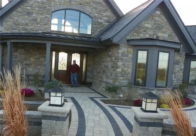 A person stands in the doorway of a stone house with a paved walkway, decorative pillars, and ornamental grasses.