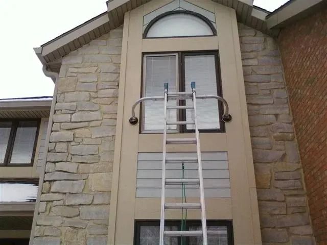 An aluminum extension ladder with a standoff stabilizer bar rests against the exterior wall of a stone house.