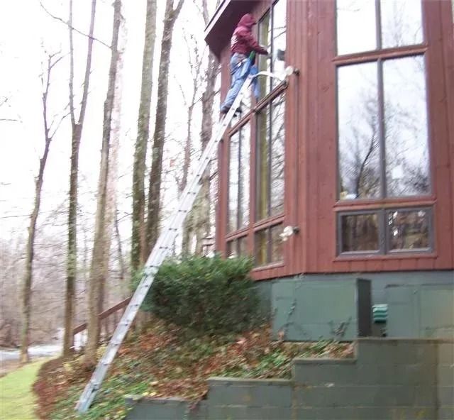 A person stands on a long ladder leaning against the second story of a brown wooden house to wash the exterior windows.