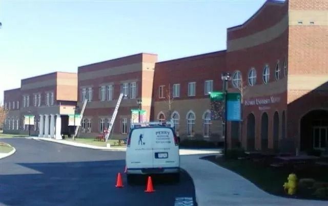A white utility van parked in front of a multi-story brick school building with a concrete walkway and orange cones.