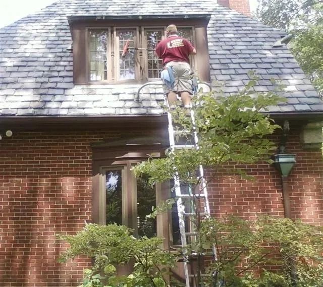 A person in a red shirt standing on a tall ladder to clean the window of a brick house with a slate roof.