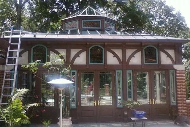 A brown timber-framed conservatory with a glass cupola, set against trees, featuring large glass doors and a tall ladder.