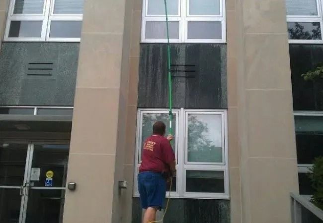 A person in a maroon shirt uses a long pole to clean the exterior windows of a tan stone building.
