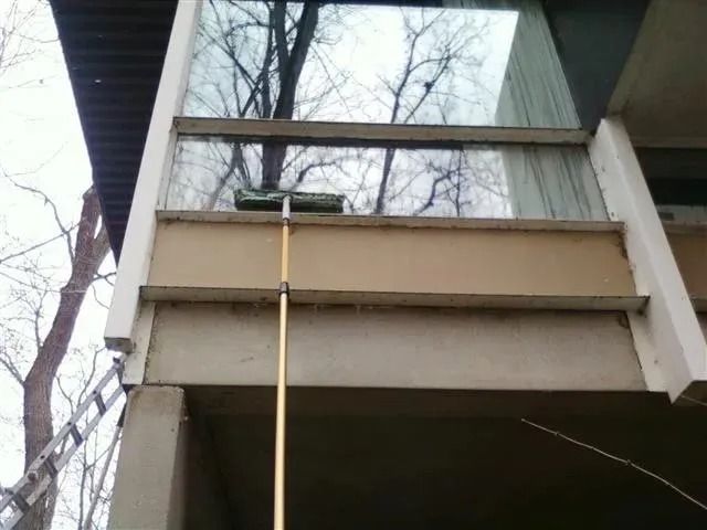 A long-handled squeegee cleans the exterior of a second-story glass window on a building with beige and white trim.