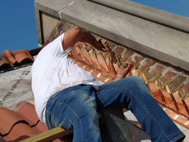 Worker crouches in rooftop gutter beside brick wall and red tiles, wearing a white shirt and jeans